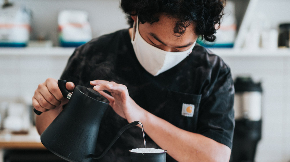 Masked cafe owner pouring water in coffee filter